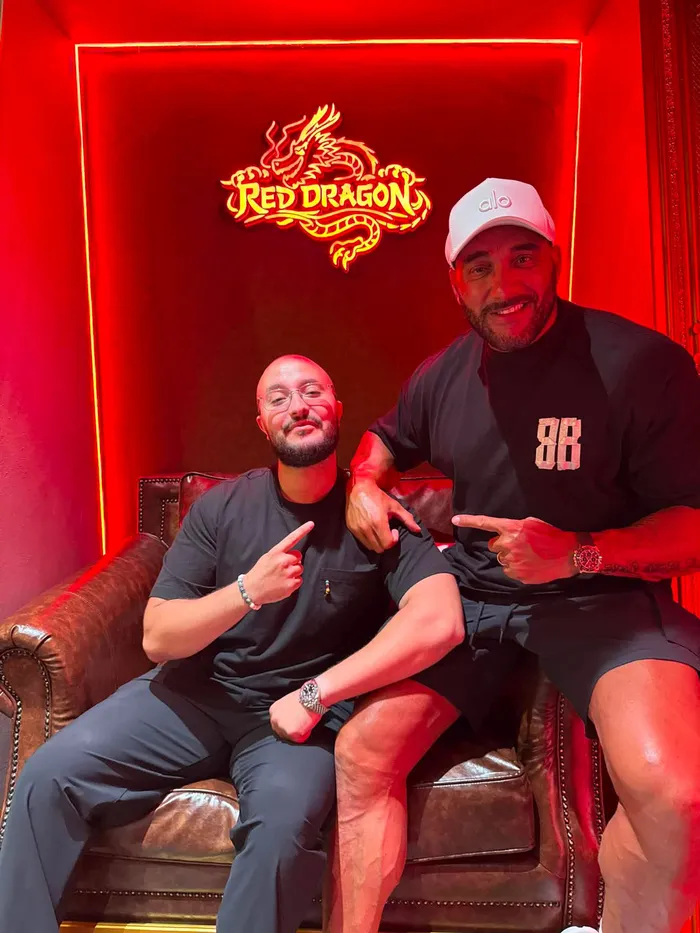 Two guests posing at the Red Dragon photo spot, one seated in the leather armchair, beneath the neon dragon emblem.